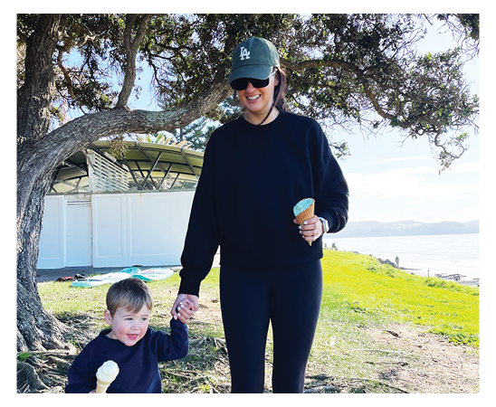 Kate White Account Director at Spruik, eating an ice cream with her son, walking outside with a tree and beach behind them.