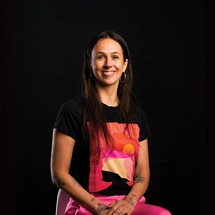 Lucie Blazevska, Senior Designer at Spruik, sitting while smiling and looking into the camera with a black background.